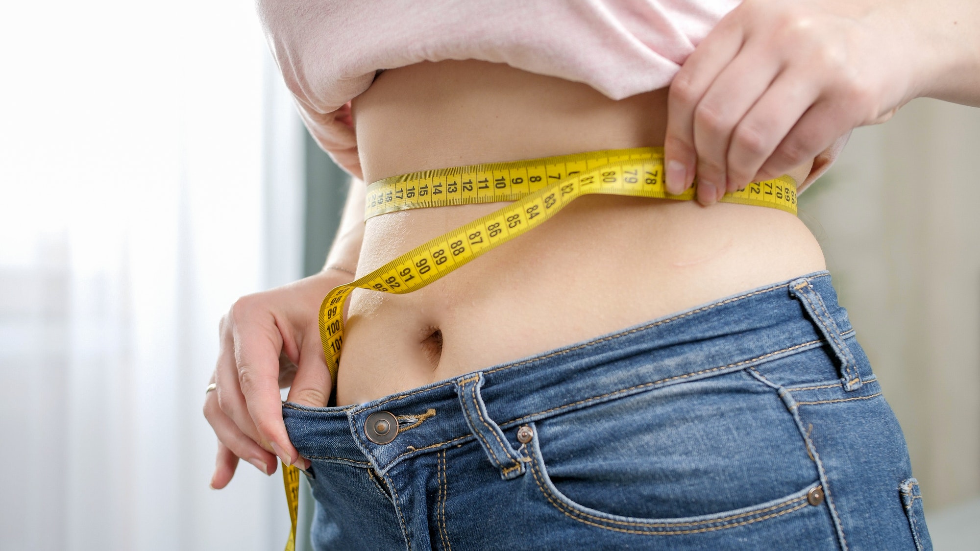 Closeup of young woman with slim belly measuring her waist at mirror in bedroom. Concept of dieting