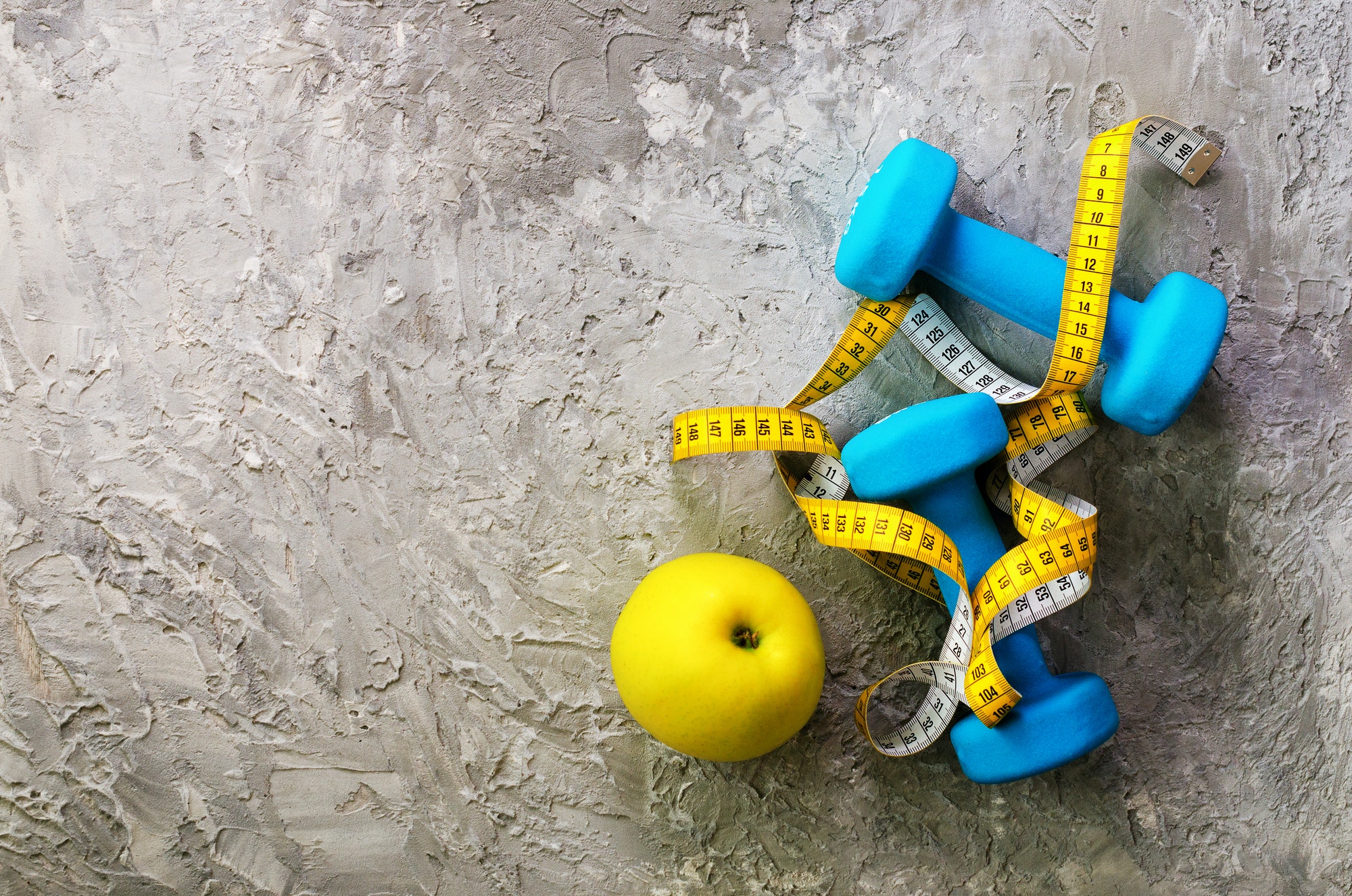 Turquoise dumbbells with measuring tape and yellow apple on concrete background. Free space for your
